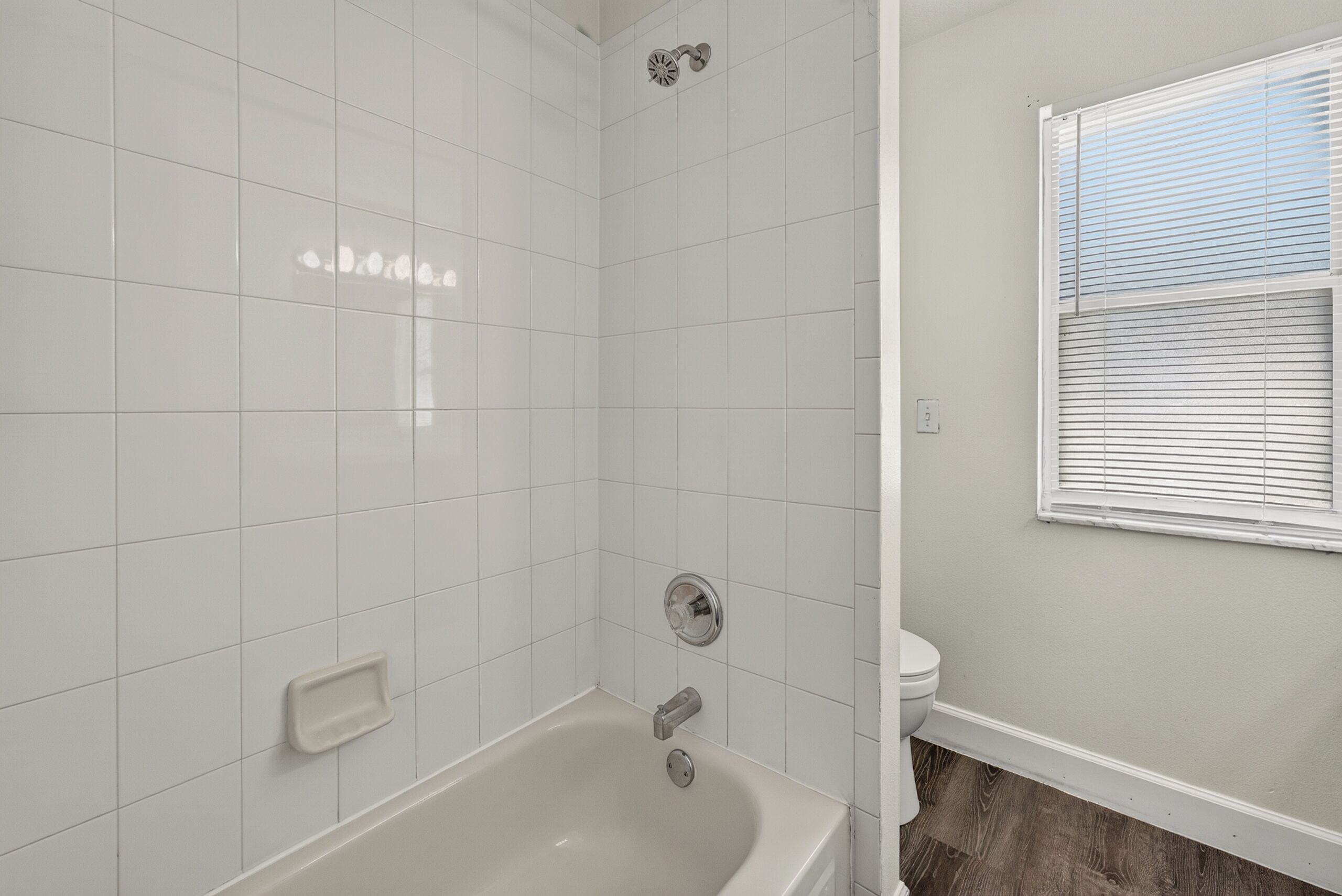 A bathroom with white tiles, a bathtub, a toilet, and a window with blinds.