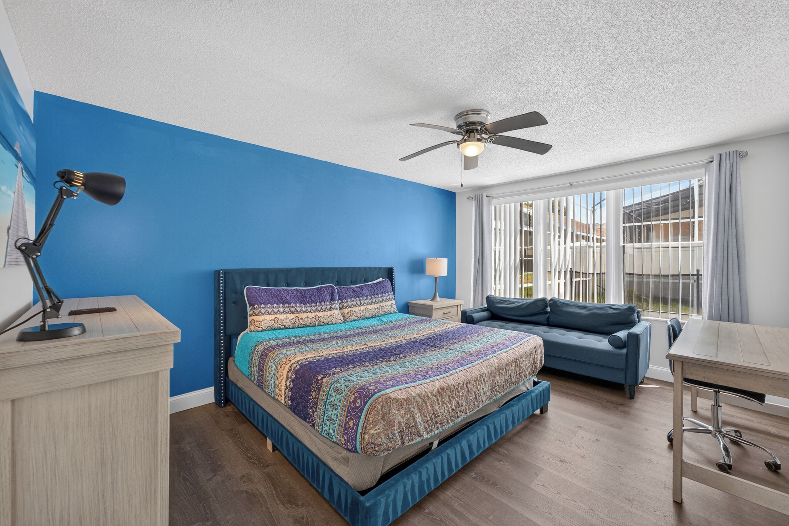 A bedroom with a bed, a desk, a chair, a ceiling fan, and a window with curtains.