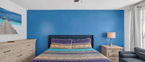 Ein Schlafzimmer mit einem Bett, einem Nachttisch, einem Schreibtisch, einem Stuhl, einem Deckenventilator und einem Fenster mit Vorhängen.