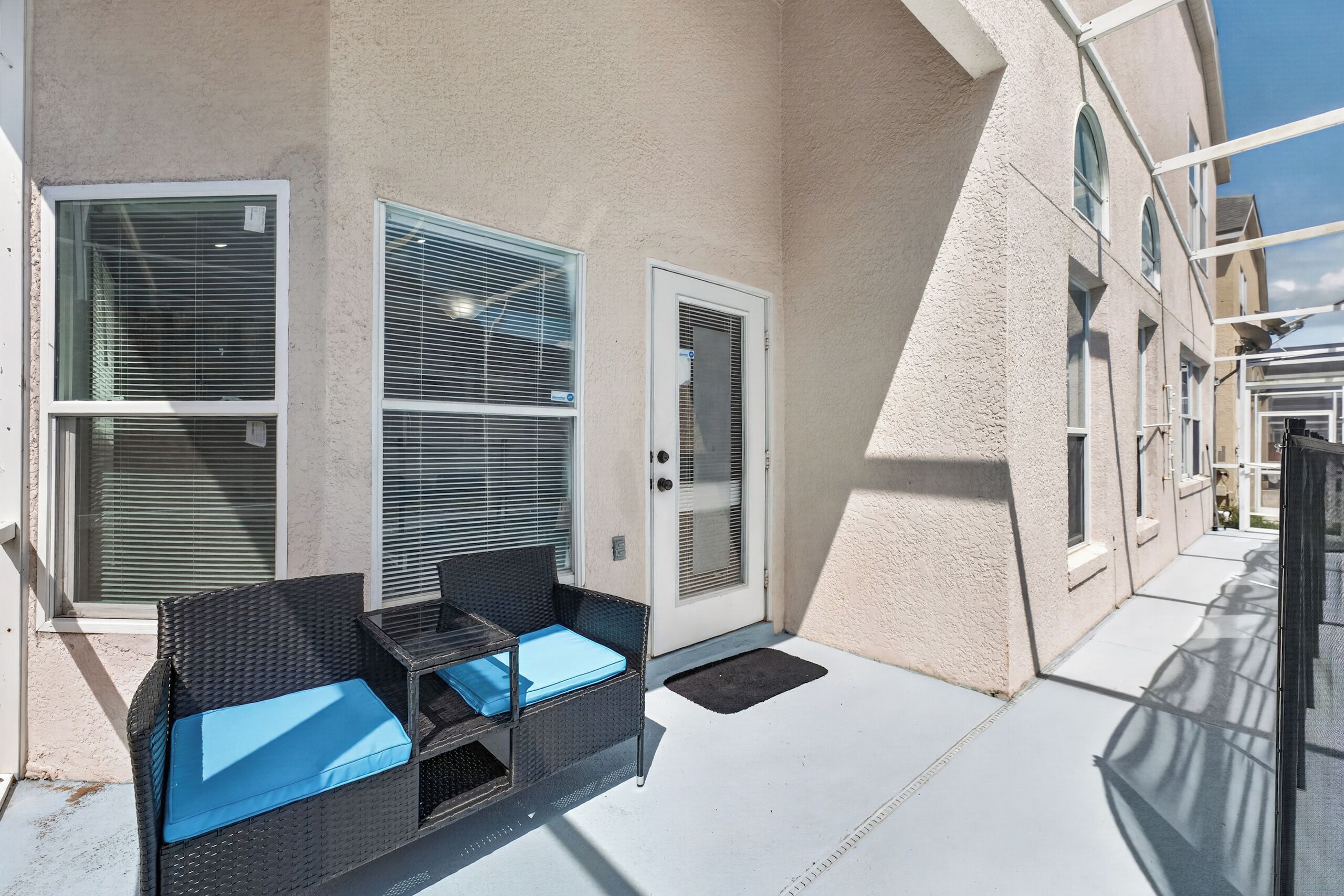 A covered outdoor seating area with a wicker table and chairs, a door, and a window with blinds.