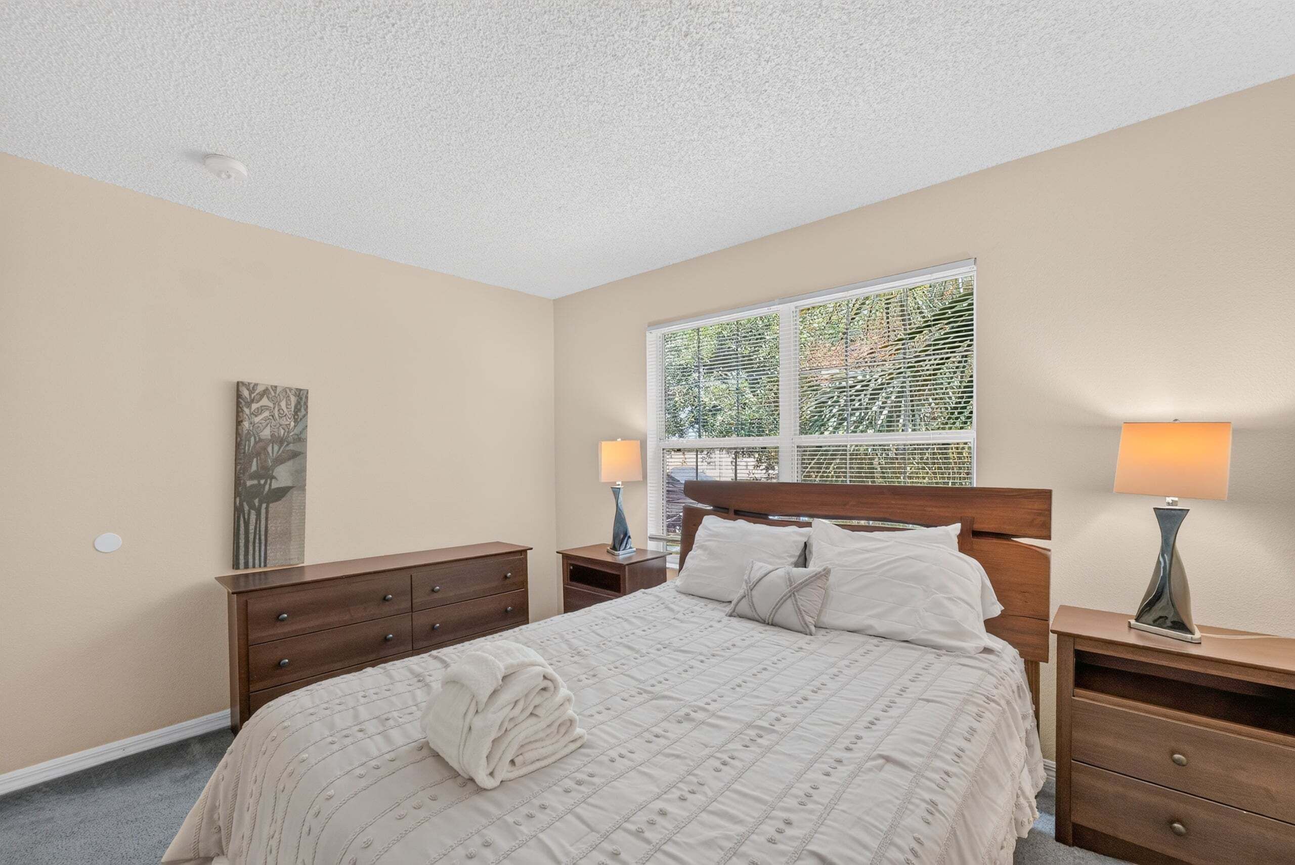 A bedroom with a bed, two nightstands, a window with blinds, and a wall-mounted lamp.
