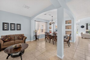 A spacious living room with a sofa, coffee table, and dining area with chairs and a chandelier. - Emerald Magic Retreat - Minutes to Disney! Emerald Island Pool Home Retreat (Kissimmee)