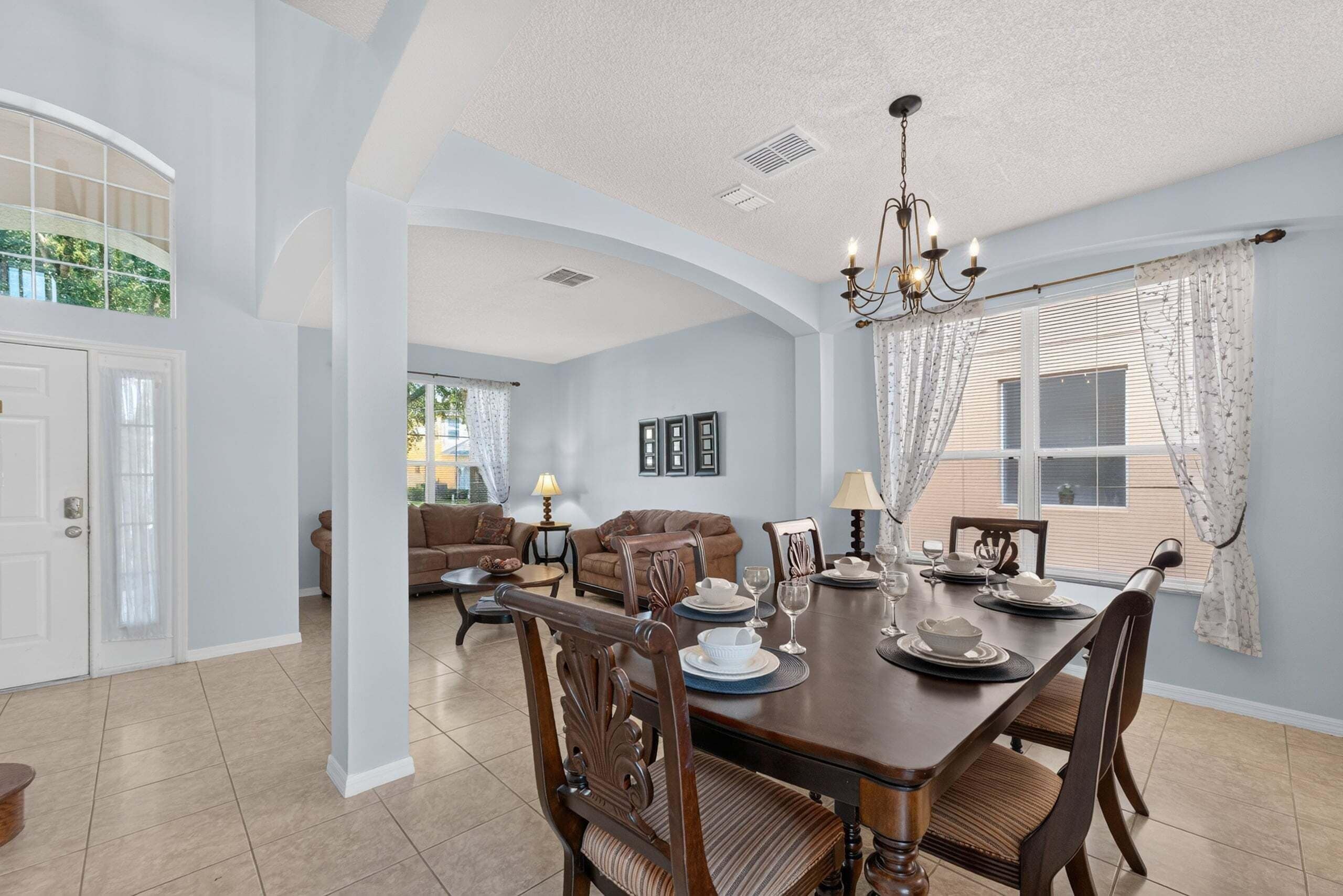 A dining area with a wooden table set for four, surrounded by chairs. There is a chandelier above the table, and the room has a high ceiling with an arched doorway leading to another room.