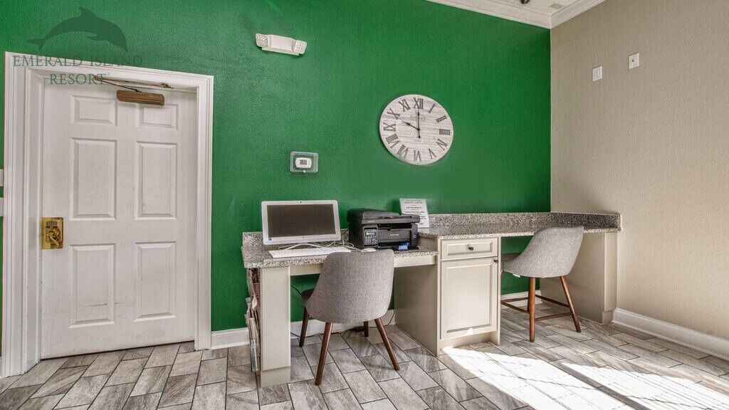 Une pièce avec un mur vert, une porte blanche, un bureau avec un ordinateur, deux chaises et une horloge.