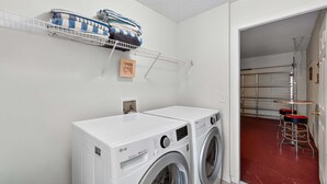 A laundry area with two front-loading washing machines, a drying rack with folded towels, and a small wooden shelf with a picture frame. - Emerald Magic Retreat - Minutes to Disney! Emerald Island Pool Home Retreat (Kissimmee)