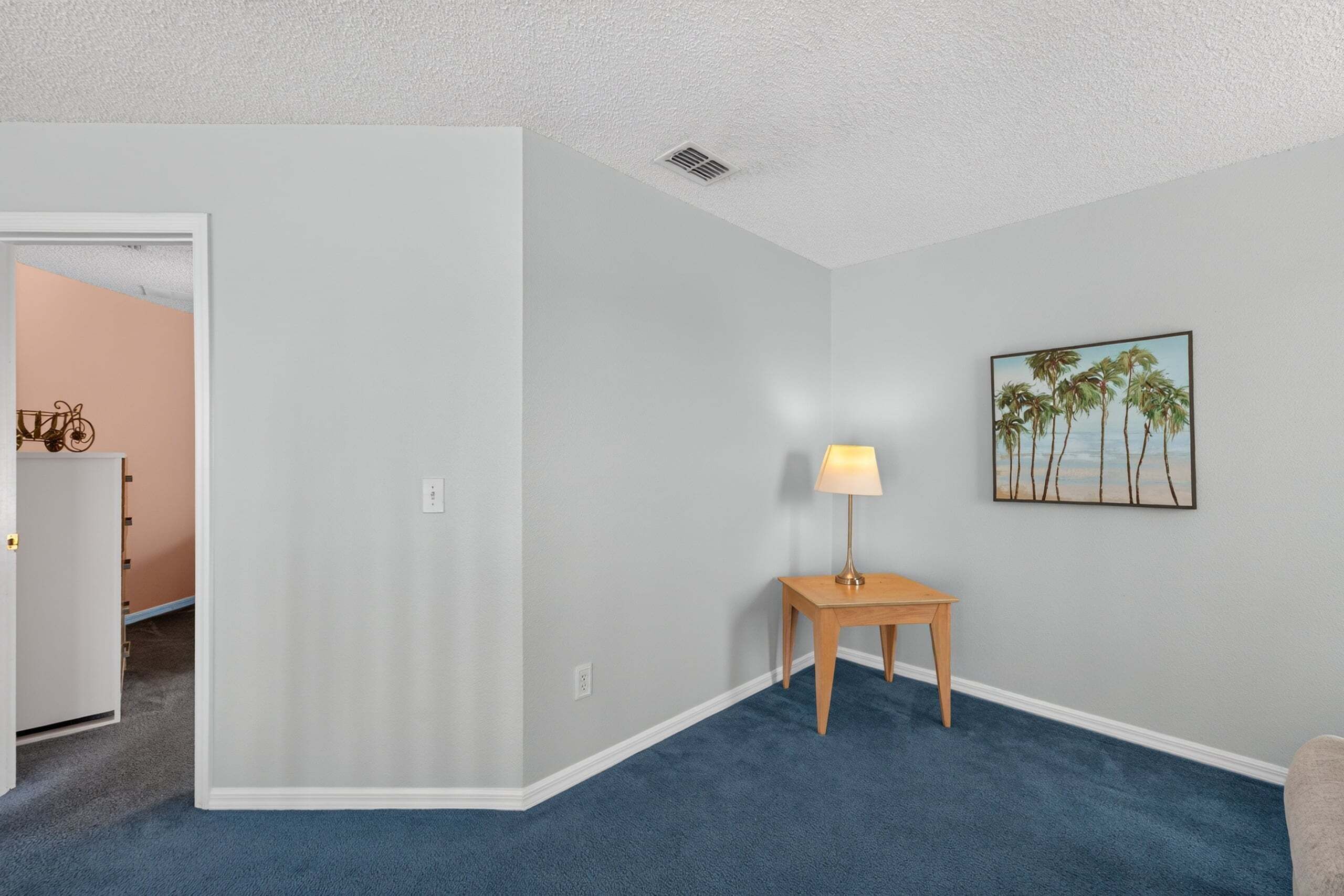 Ein Zimmer mit blauem Teppich, einem Holznachttisch mit Lampe und einem an der Wand hängenden Bild von Palmen.