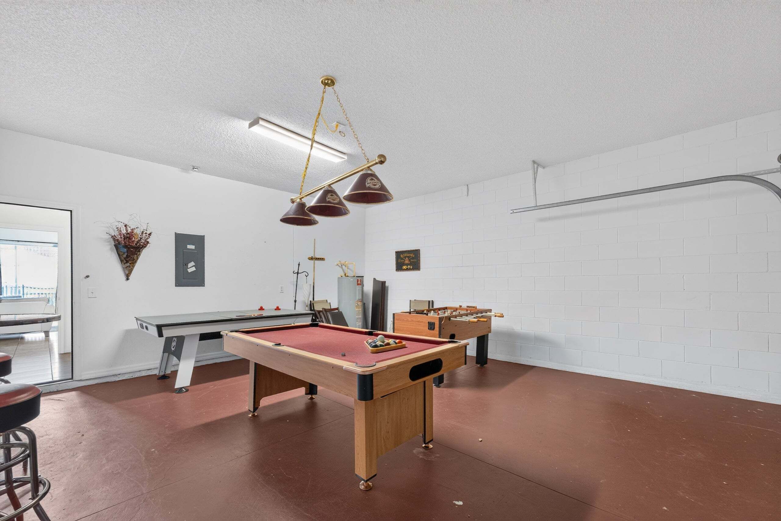 A pool table, a foosball table, and a bar stool in a room with white brick walls and a ceiling light fixture.