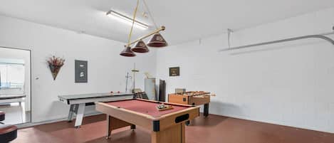 Ein Billardtisch, ein Kicker und ein Barhocker in einem Raum mit weißen Ziegelwänden und einer Deckenleuchte.