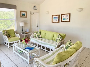 A room with a white wicker sofa set, a glass coffee table with books, a lamp, and framed artwork on the wall. - The Anchorage | Steps from Rainbow Bay Beach, Eleuthera (North Eleuthera)