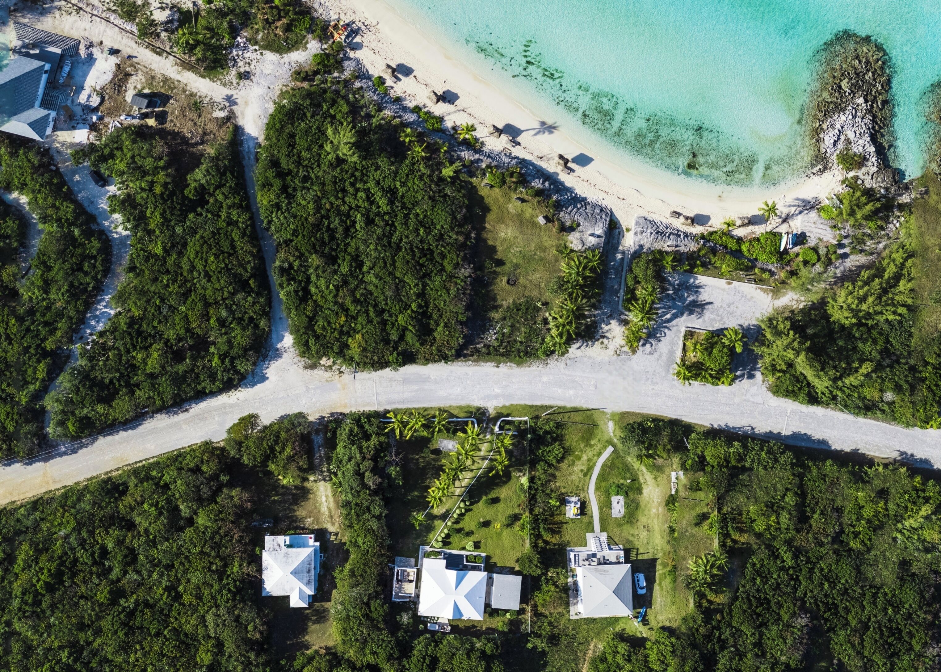 Vue aérienne d’une propriété située sur la côte, avec une plage, une végétation luxuriante et plusieurs bâtiments dotés de toits plats.