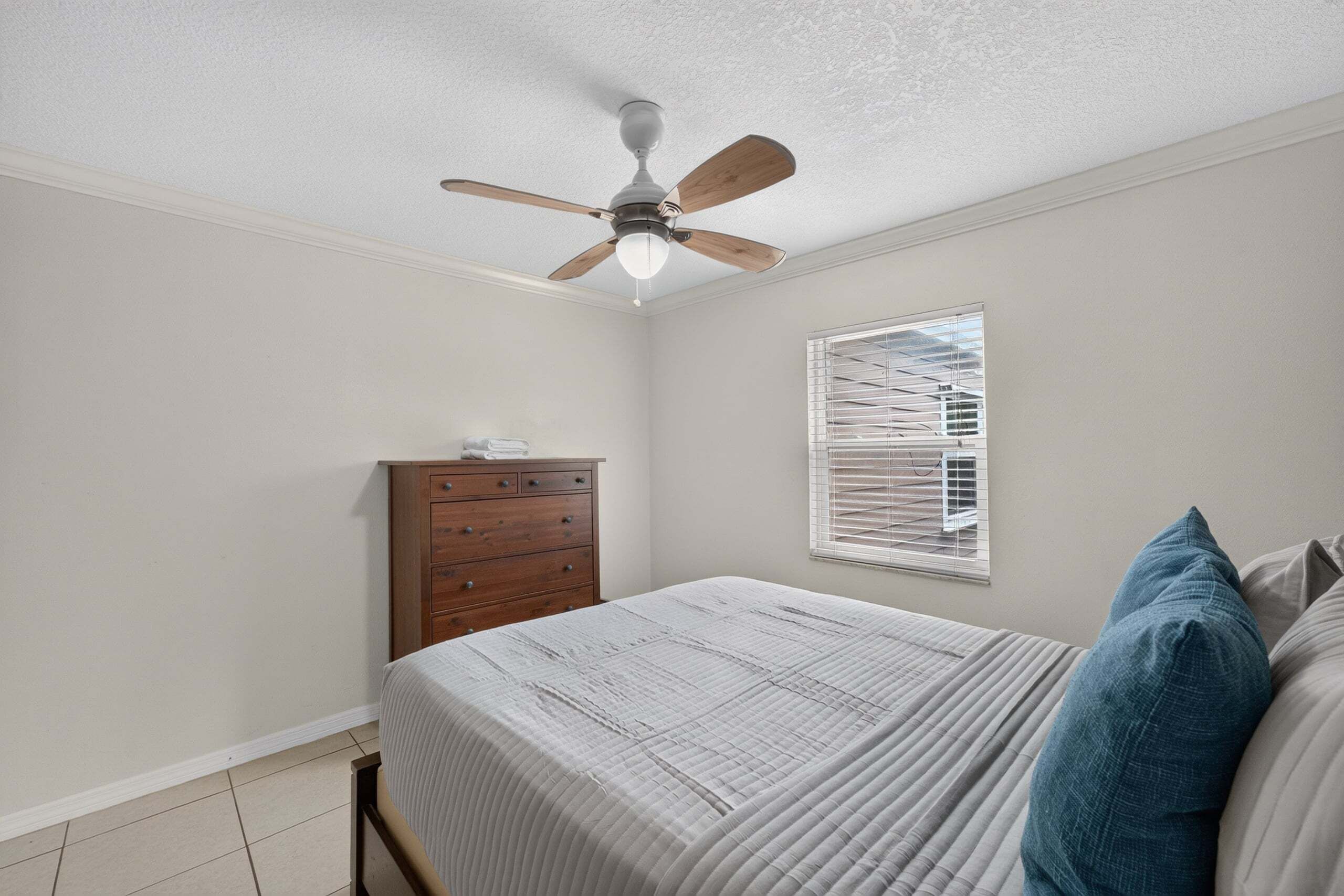 A bedroom with a bed, a wooden dresser, a ceiling fan, and a window with blinds.