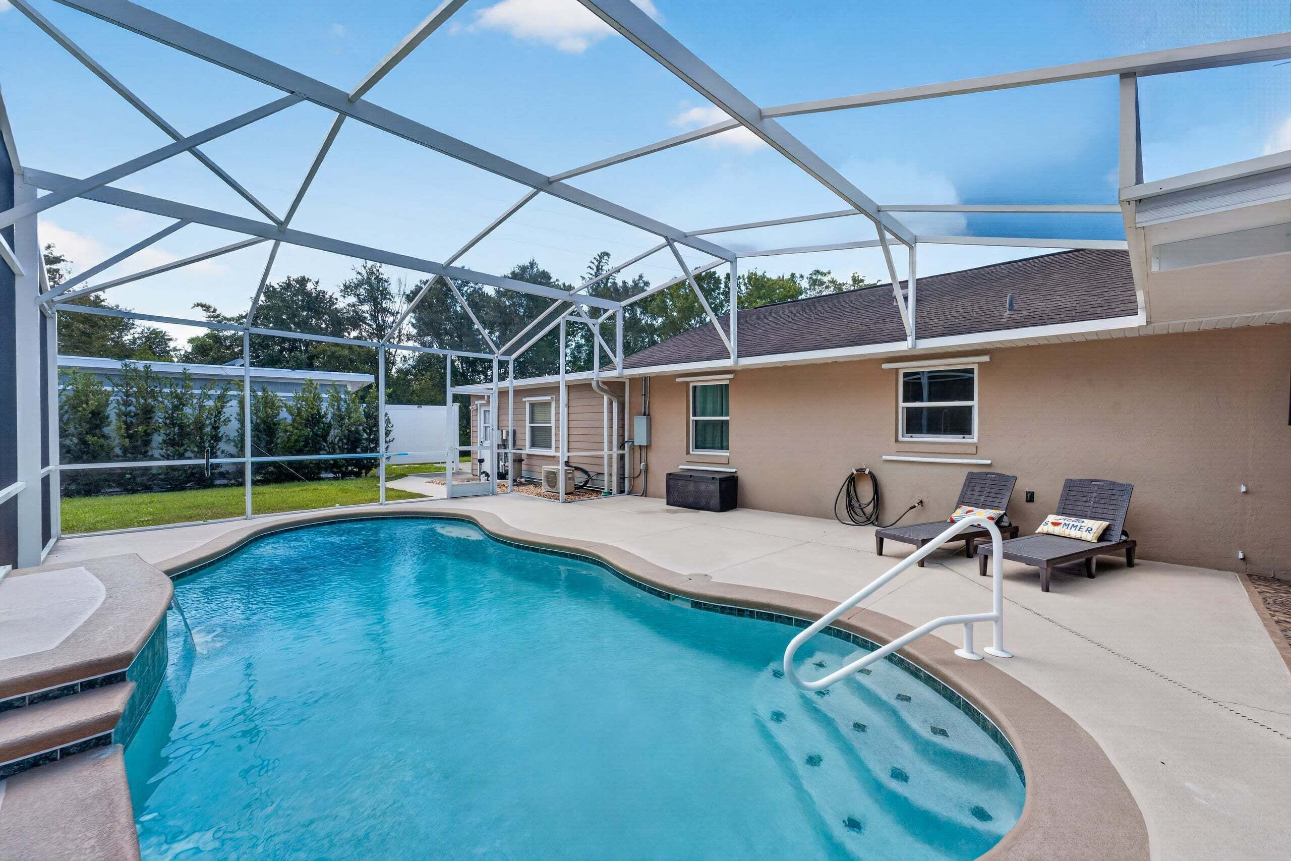 A covered outdoor swimming pool with a clear sky, a house in the background, and a patio area with lounge chairs.