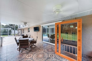 Une terrasse couverte comprenant un espace repas, un ventilateur de plafond et une vue sur le jardin arrière, accessible par des portes vitrées.