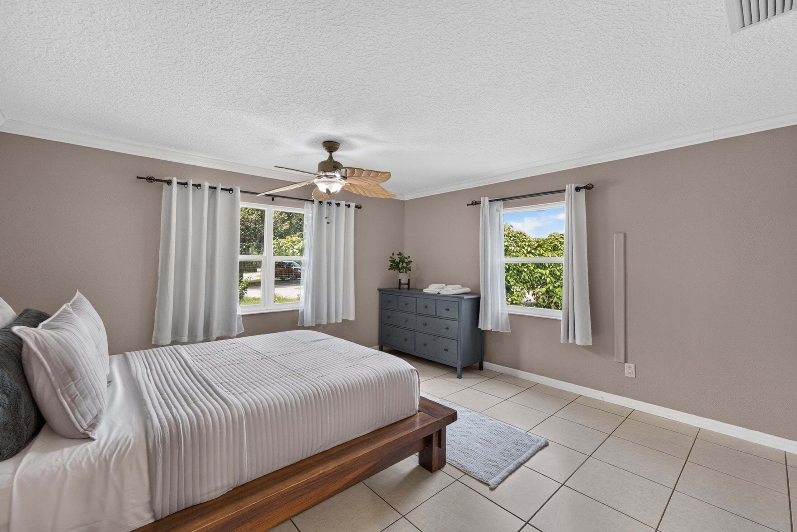 Quarto com cama, cômoda, ventoinha no teto e janela com cortinas.