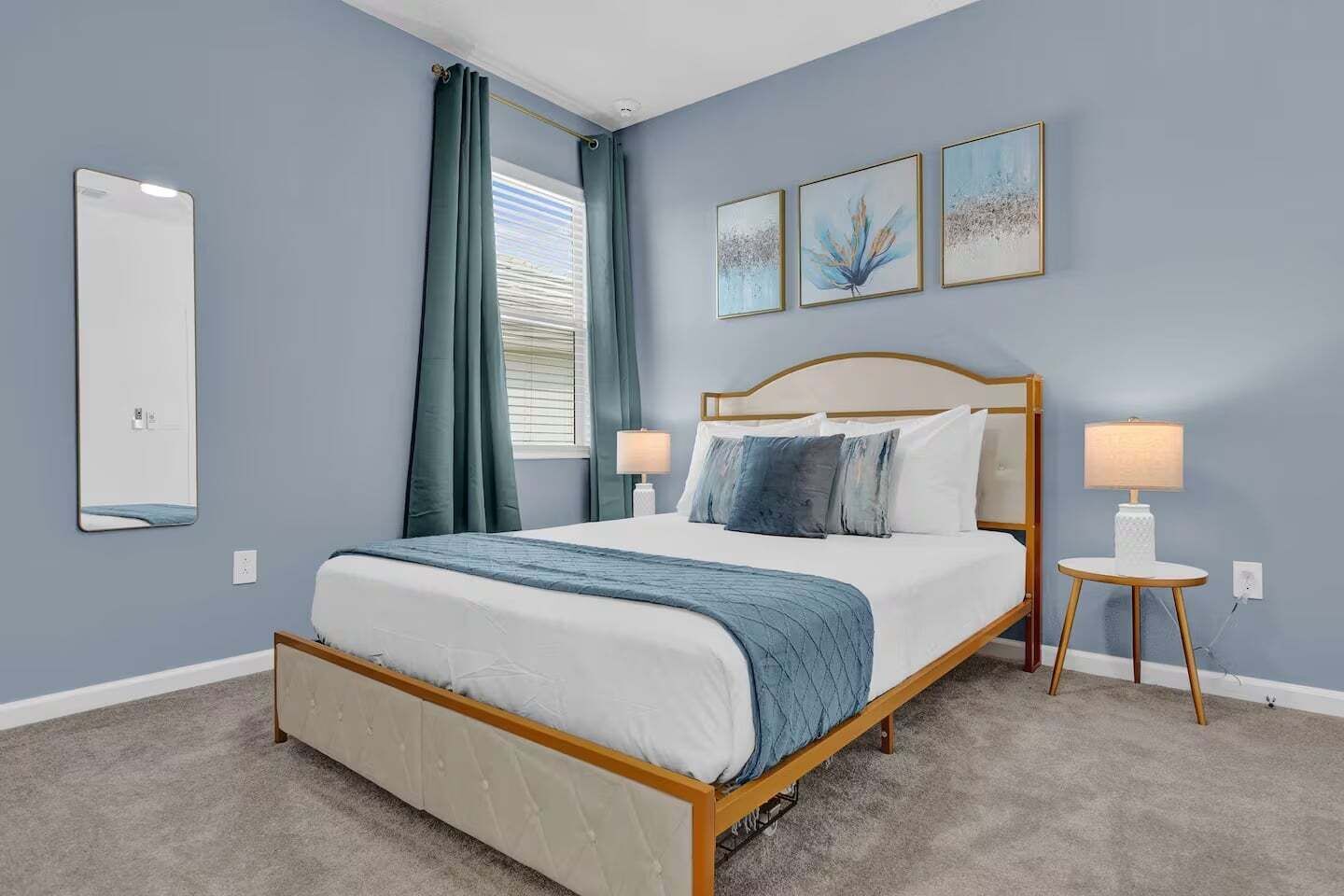 A bedroom with a bed, two bedside lamps, a window with curtains, and three wall-mounted artworks.