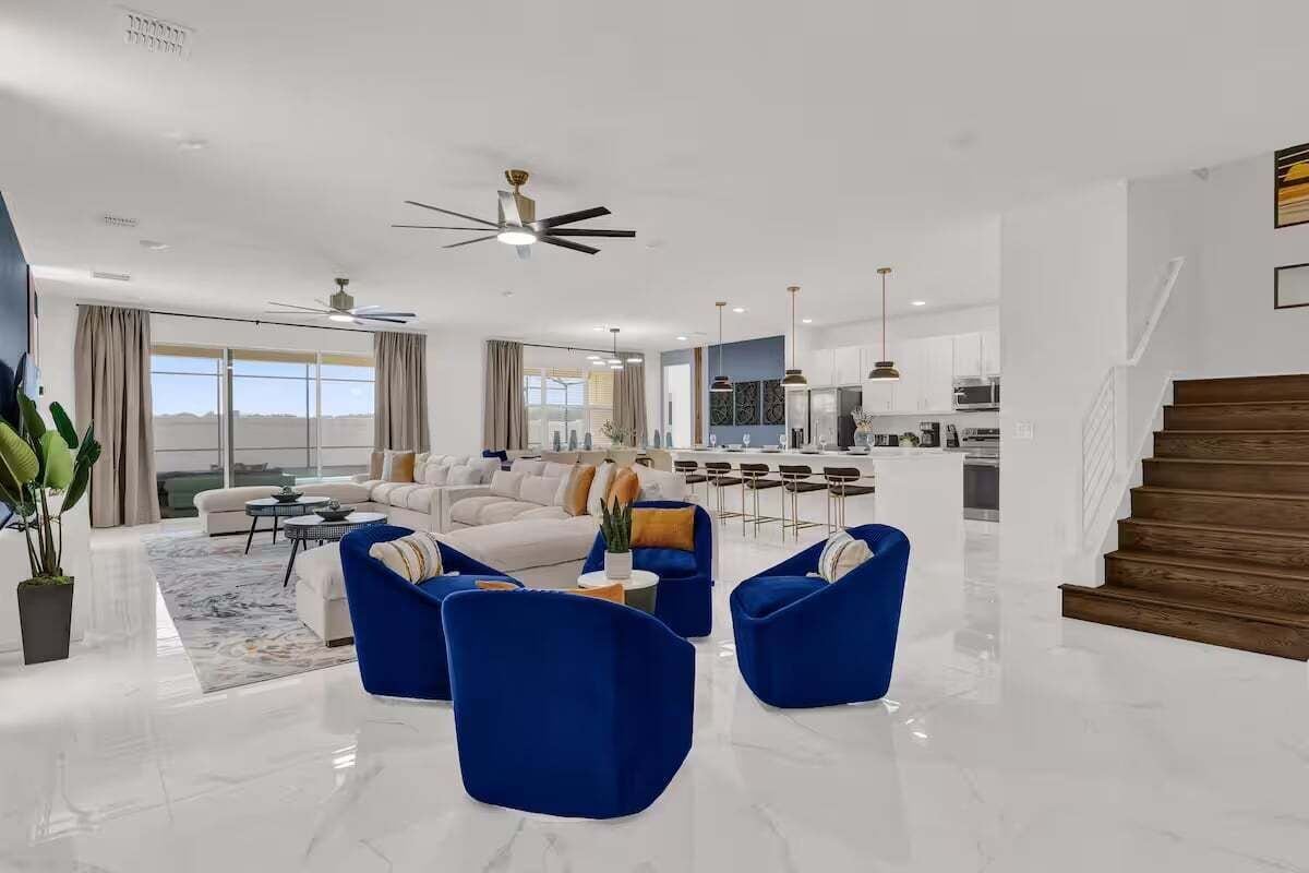 A spacious living area with a modern design, featuring a large sofa set, blue accent chairs, and a kitchen in the background.