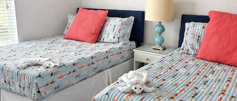 Un dormitorio con dos camas, cada una con un edredón estampado, almohadas rojas y una manta blanca. Hay una mesita de noche con una lámpara, un cuadro en la pared y una ventana con persianas.