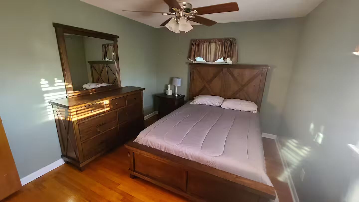 A bedroom with a wooden bed, a dresser, a nightstand, a ceiling fan, and a window with curtains.
