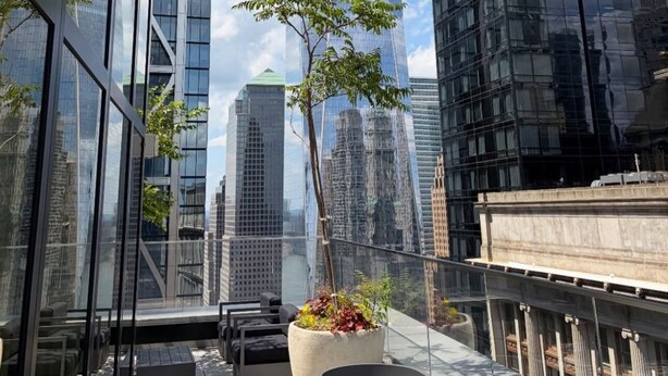 A modern building with a glass facade, surrounded by other high-rise structures, and a rooftop terrace with outdoor furniture and a potted plant.