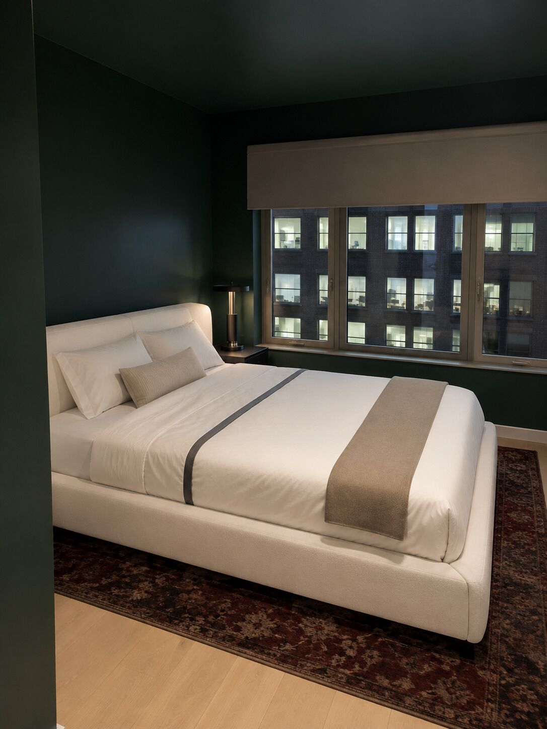 A modern bedroom with a large bed, a nightstand, a lamp, and a view of a building through a window.