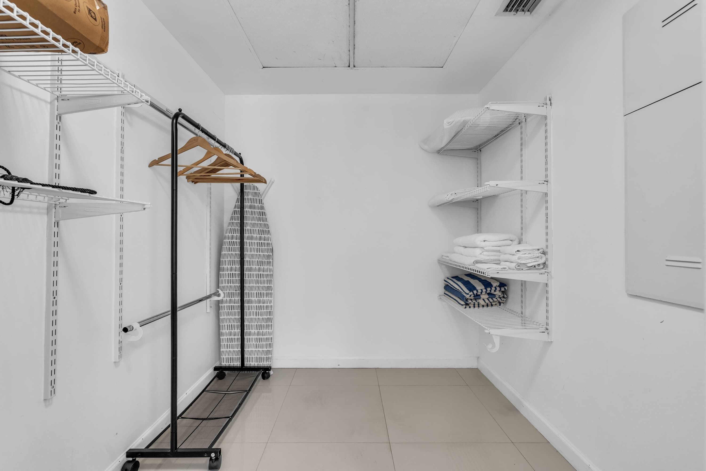 A compact, white-walled walk-in closet with metal shelving units, a clothing rack, and a hanging garment bag.