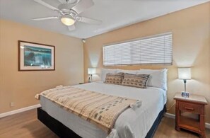A bedroom with a bed, a ceiling fan, a window with blinds, a bedside table with a lamp, and a framed picture on the wall. - HUGE SALE!! OCEAN FRONT and BEACH FRONT . Ground floor just steps from the Ocean (Kihei)