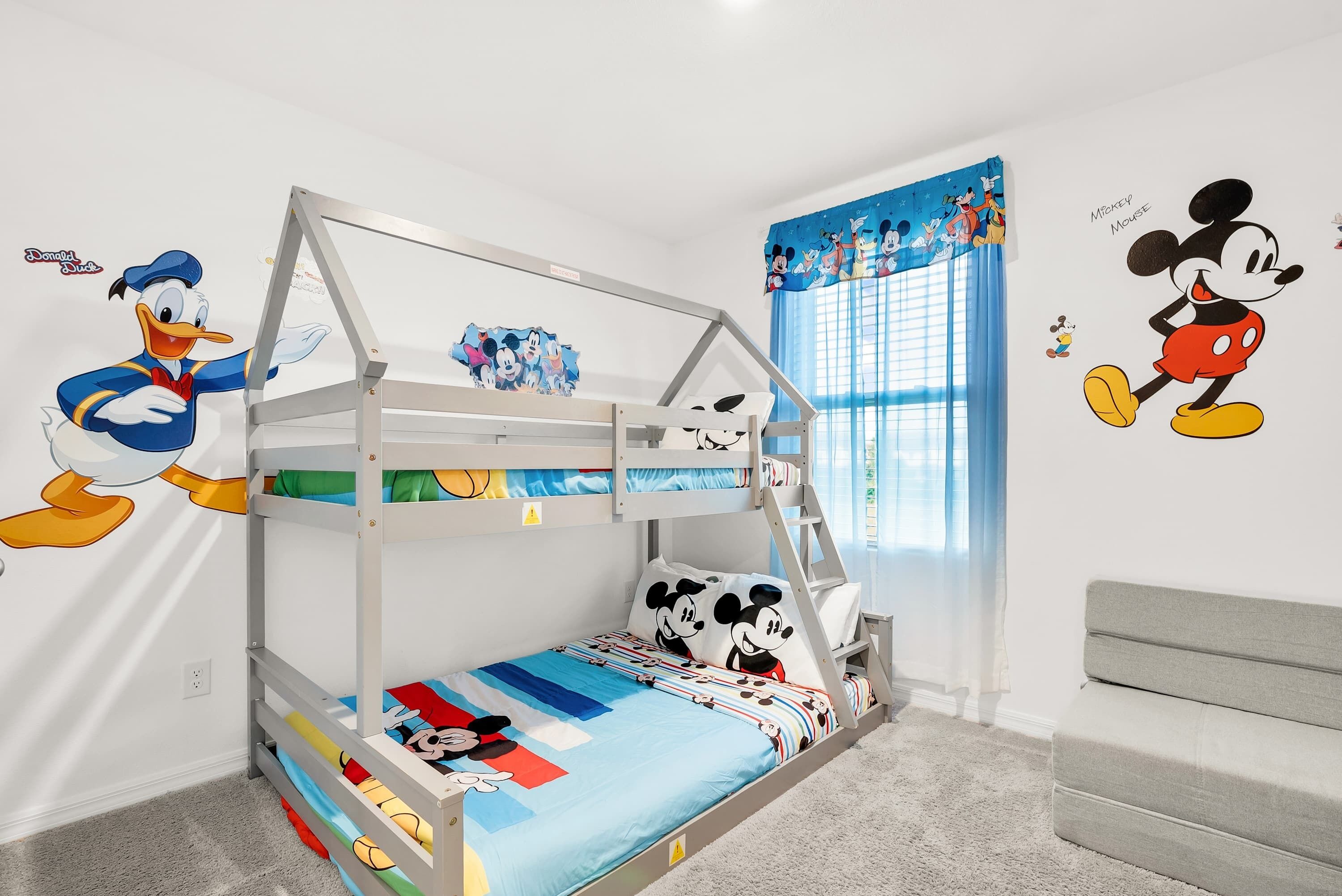 Un lit superposé décoré d’autocollants représentant Mickey Mouse et Donald Duck, une fenêtre avec des rideaux bleus et un canapé gris.