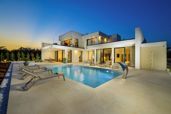 Una casa moderna de dos pisos con piscina, mobiliario exterior y un patio bien iluminado.