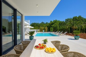 Un área de piscina exterior moderna con sillones de descanso y una mesa de comedor con frutas y verduras.