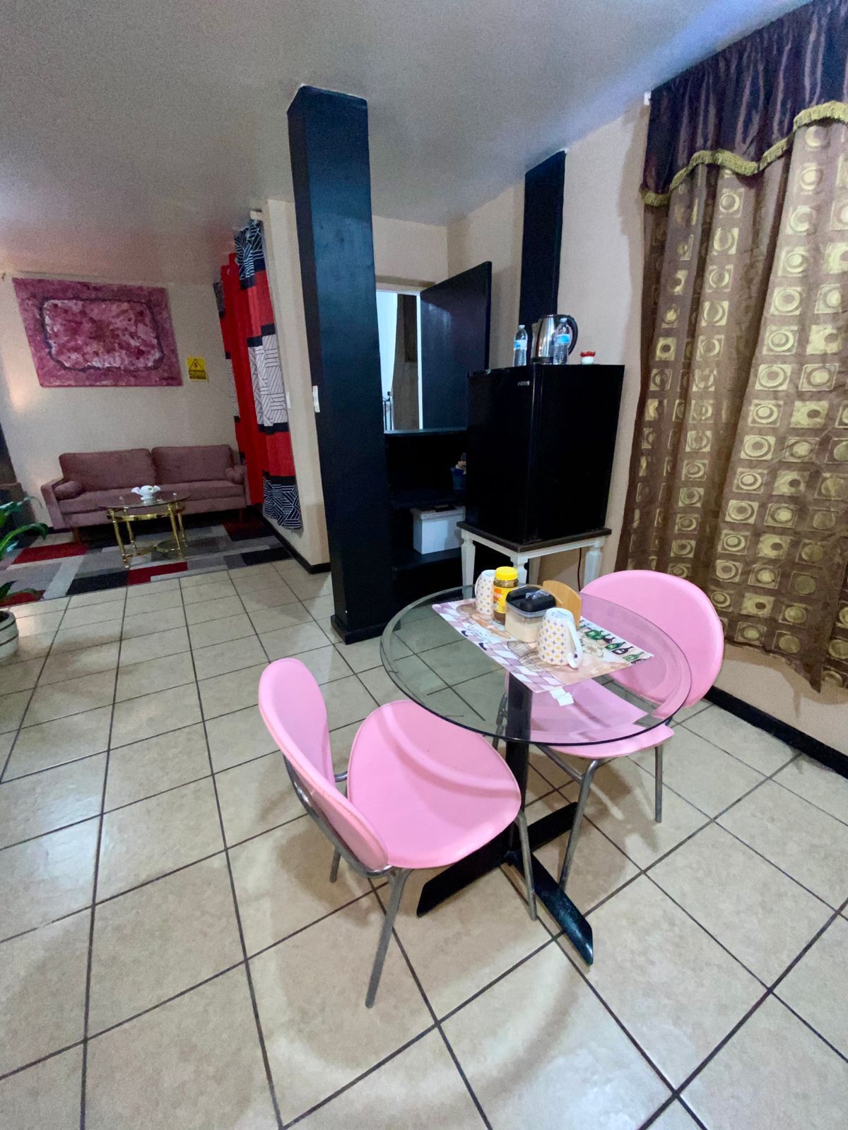 A living room with a sofa, a glass-top dining table with pink chairs, a flat-screen TV, and a patterned curtain.