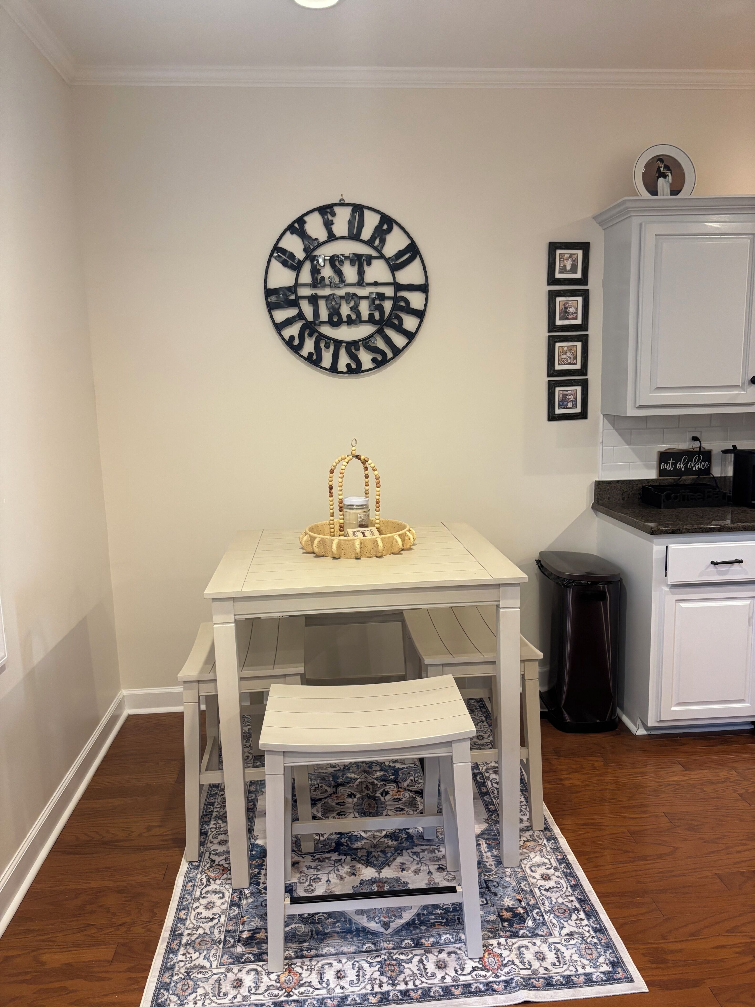 A kitchen with a white table, a patterned rug, a wall clock, and framed pictures.