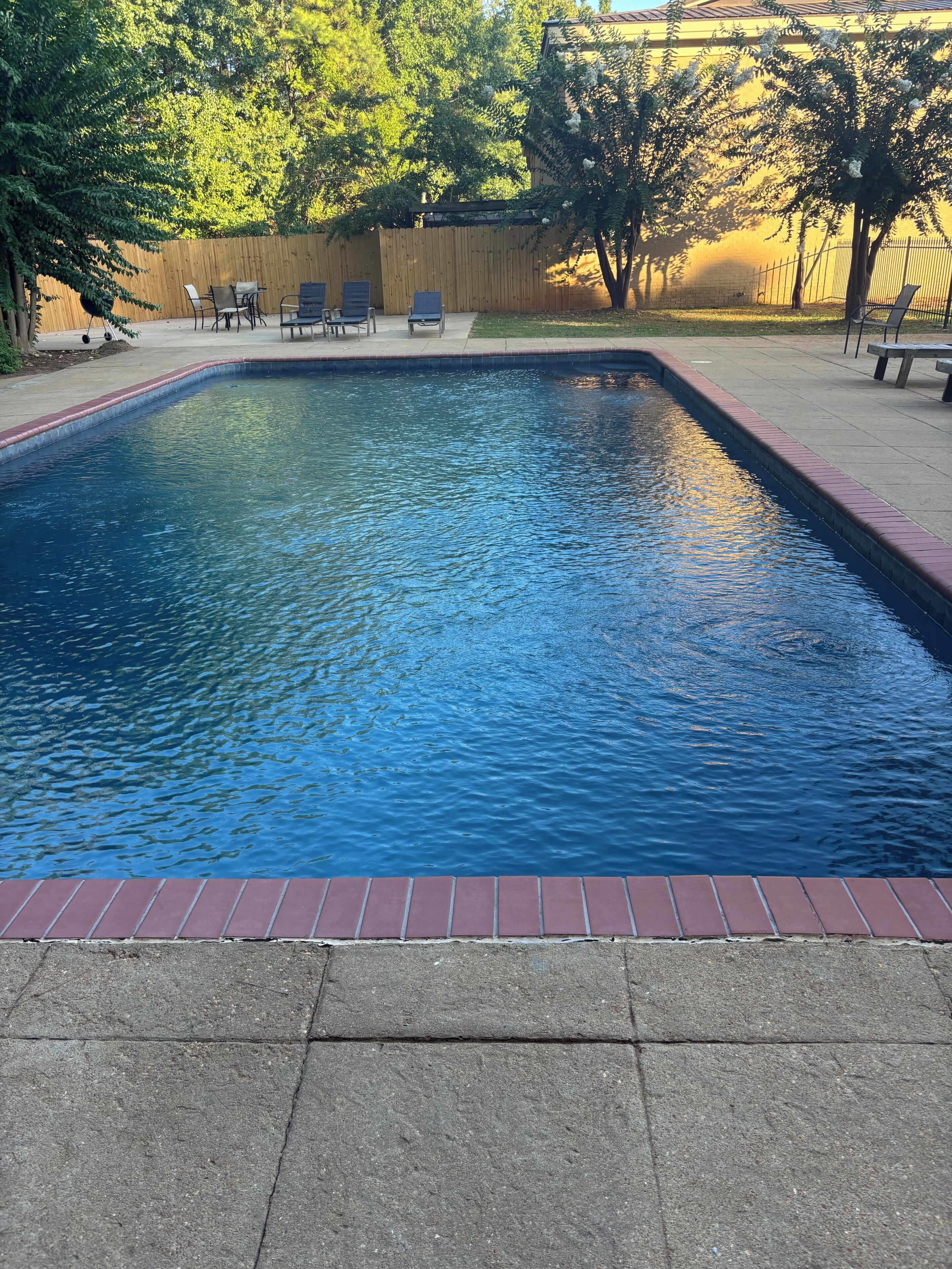 A rectangular swimming pool with a brick edge, surrounded by a wooden fence and trees.
