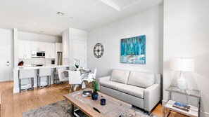 A modern living room with a grey sofa, a coffee table, and a dining area with white cabinets and a kitchen in the background. - The Perfect Old Naples Location; Walk to 5th Ave & the Beach! (Naples)