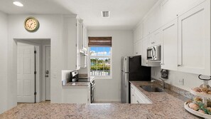 A kitchen with white cabinets, a microwave, a refrigerator, a sink, and a clock on the wall.