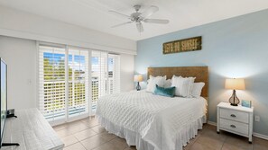 Une chambre à coucher comprenant un lit, une table de chevet, une lampe, un ventilateur de plafond et une vue sur l’extérieur à travers une fenêtre munie de stores.