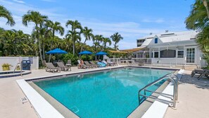 A clear blue pool with lounge chairs and umbrellas, surrounded by palm trees and a building with a white facade.