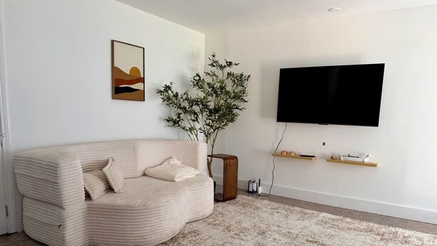 Una sala de estar con un sofá color beige, un televisor de pantalla plana grande montado en la pared, una planta en maceta y una mesita de madera.