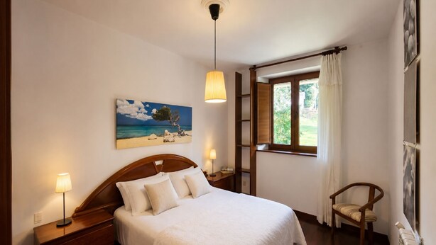 A bedroom with a bed, bedside lamps, a painting of a beach scene, a window with curtains, and a chair. - El Joyu Erías | House with a fenced-in garden in Llanes (Vidiago)