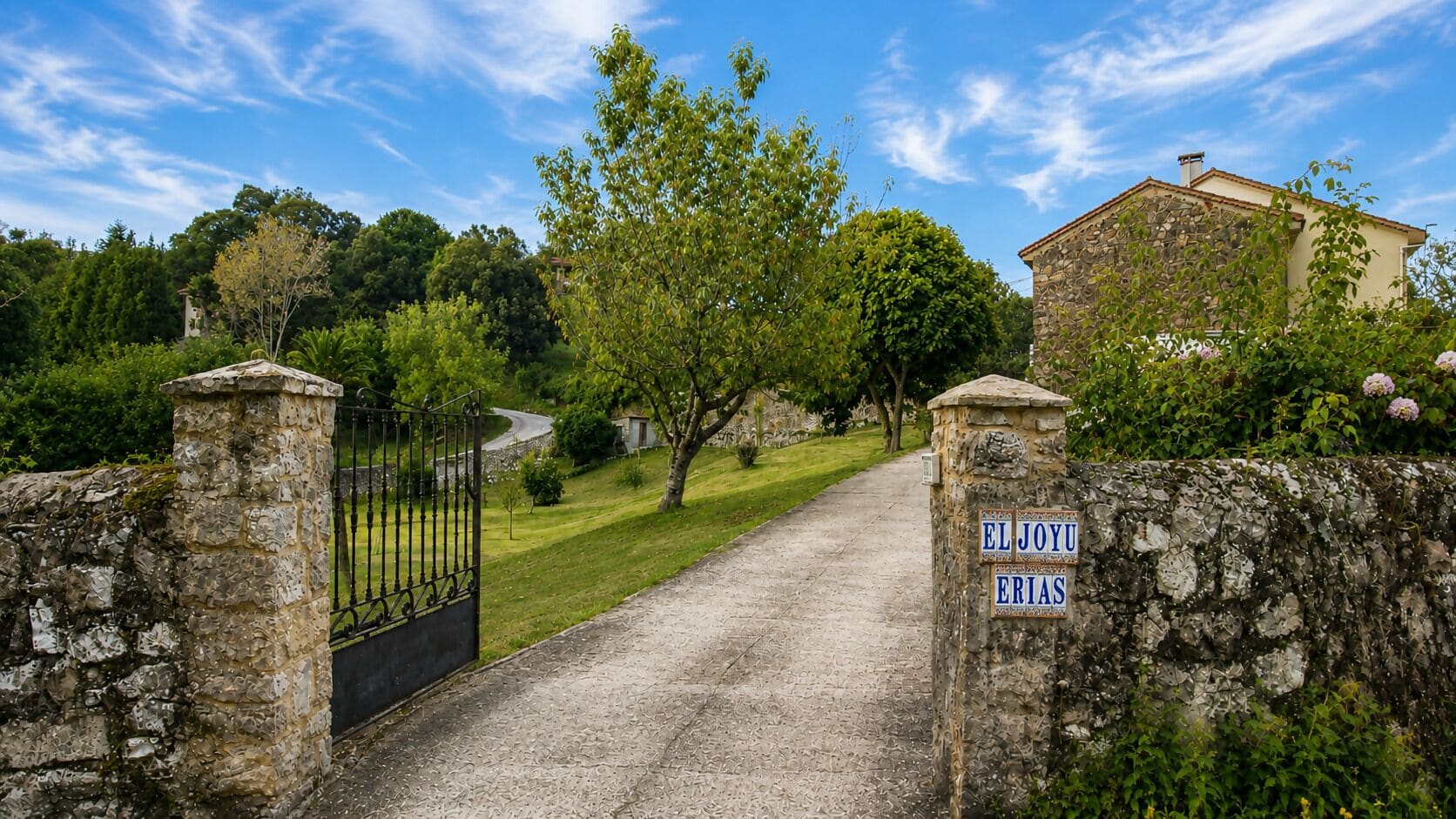 A stone gate with a metal gate, leading to a pathway flanked by greenery and a stone wall.