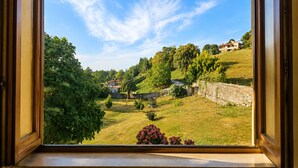 A view through a window of a grassy area with trees and a stone wall, with buildings in the background. - El Joyu Erías | House with a fenced-in garden in Llanes (Vidiago)