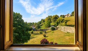 A view through a window of a grassy area with trees and a stone wall, with buildings in the background. - El Joyu Erías | House with a fenced-in garden in Llanes (Vidiago)