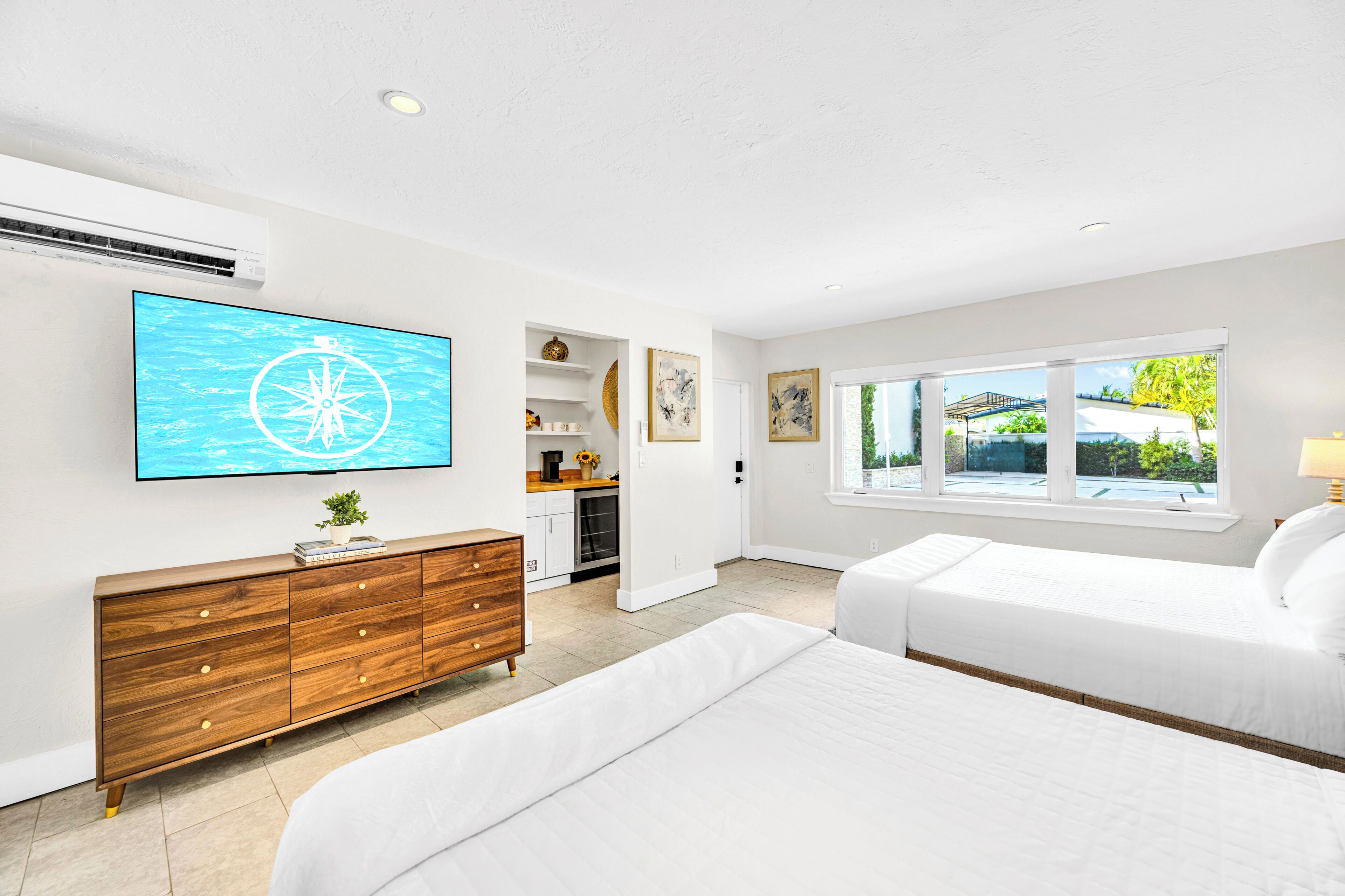 Une chambre moderne avec un grand téléviseur, une commode en bois, deux lits et une vue sur l’extérieur par la fenêtre.