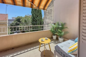 Ein Balkon mit einem gelben Tisch, zwei Blumentöpfen, einem weißen Geländer und einer Couch mit einem gestreiften Kissen.