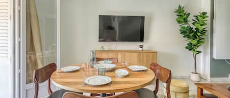Un comedor con una mesa redonda de madera dispuesta para cuatro personas, rodeada de sillas de madera con asientos acolchados. Un televisor está montado en la pared sobre la mesa.