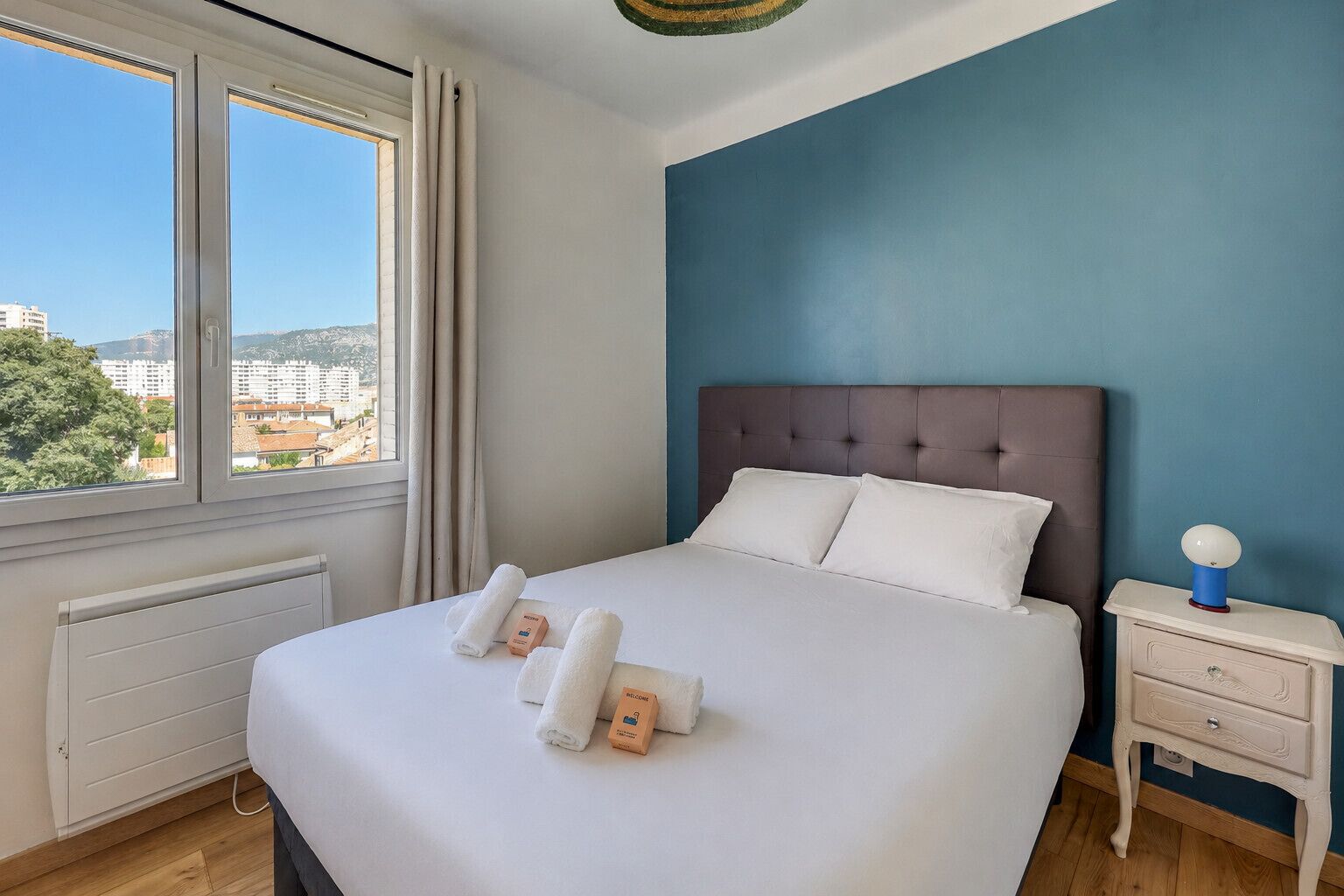 A hotel room with a large bed, a nightstand, a window with curtains, and a view of buildings outside.