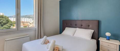 A hotel room with a large bed, a nightstand, a window with curtains, and a view of buildings outside.