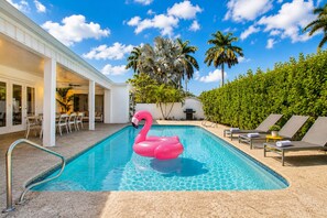 Piscina con un flotador de flamenco rosa, sillas de descanso, palmeras y un área de patio techada.