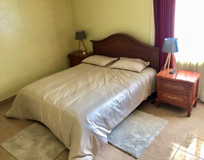 Ein ordentlich bezogenes Bett mit weißer Daunendecke, zwei Nachttische mit Lampen, ein Holzbeistelltisch und ein heller Teppich auf Fliesen.