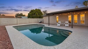 Una piscina con superficie de agua azul cristalina, rodeada por un área ajardinada con un muro de ladrillo y un patio cubierto con sillas y una mesa.