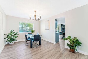 Ein moderner Essbereich mit einem Glastisch, schwarzen Stühlen und einer Chandelieber. Üchtöpfe und ein Fenster mit Jalousien sind ebenfalls zu sehen.